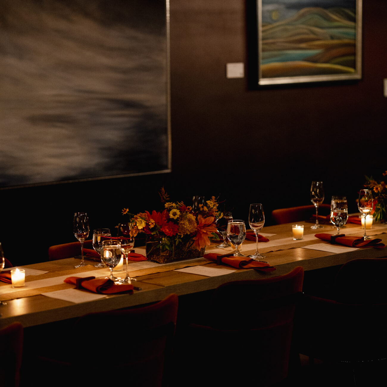 A long dining table set with candles, flowers, wine glasses, and napkins in a dimly lit room with artwork on the walls.